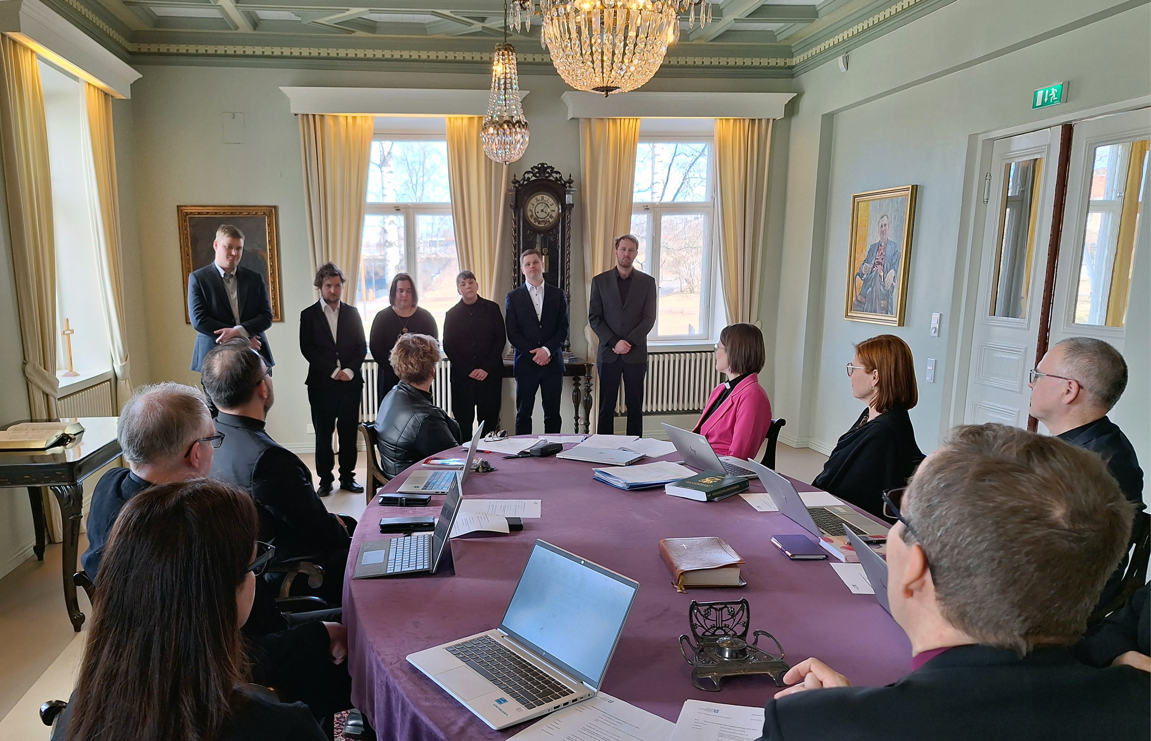 Ihmiset pöydän ympärillä katselevat rivissä seisovia kuutta henkilöä, jotka saavat pian pappisvihkimyksen.